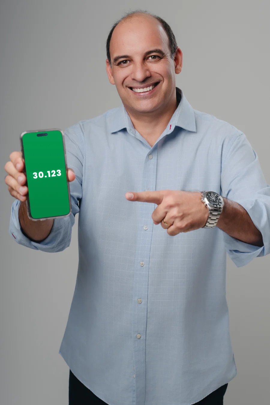 Ari Saraiva de camisa azul-clara segurando um celular com a etiqueta do número 30.123 na tela e apontando para ele.